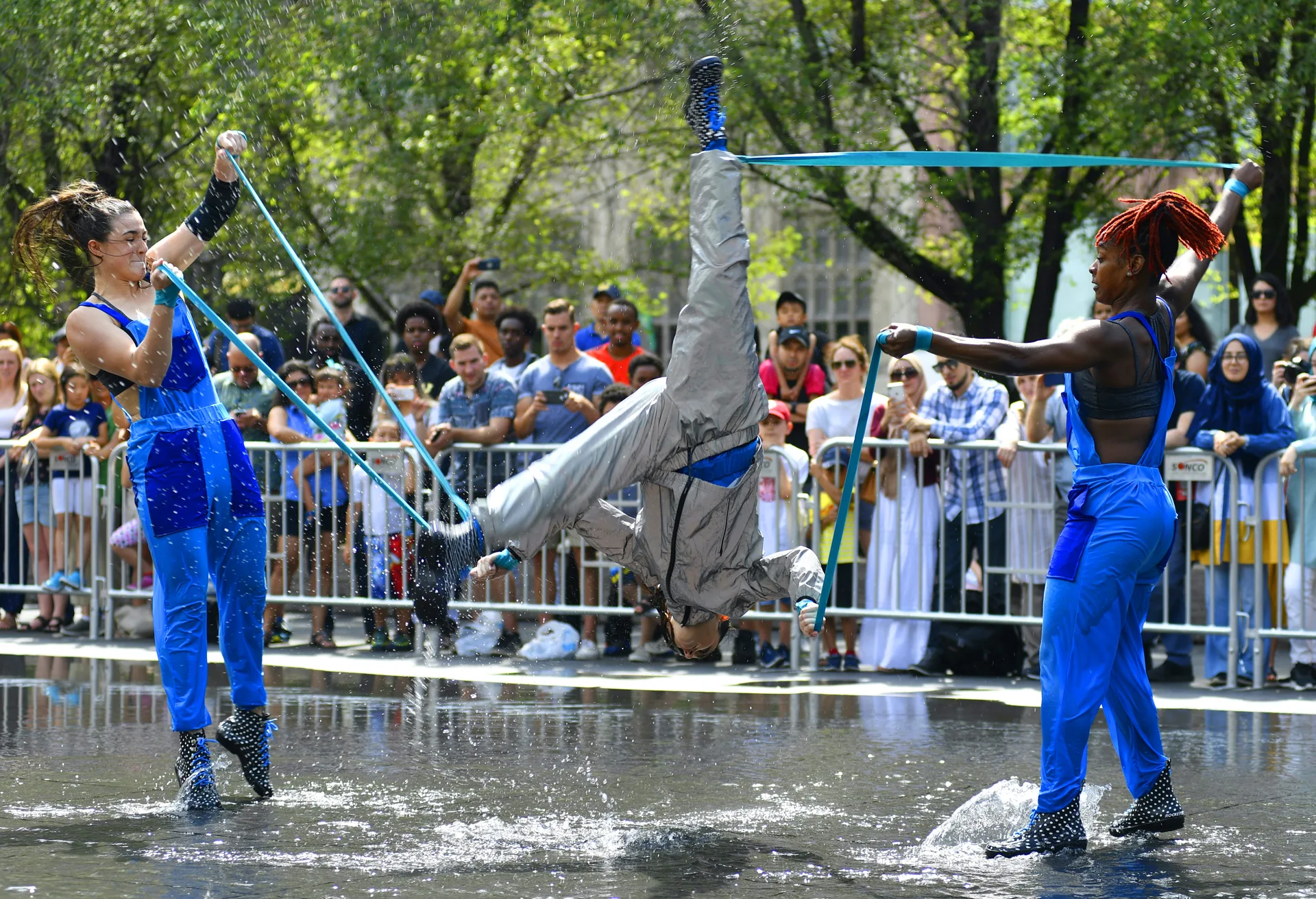 Three dancers perform outdoors in front of a crowd. They are standing in a shallow fountain. Two dancers wearing blue coveralls are supporting a third dancer doing a flip with blue straps.