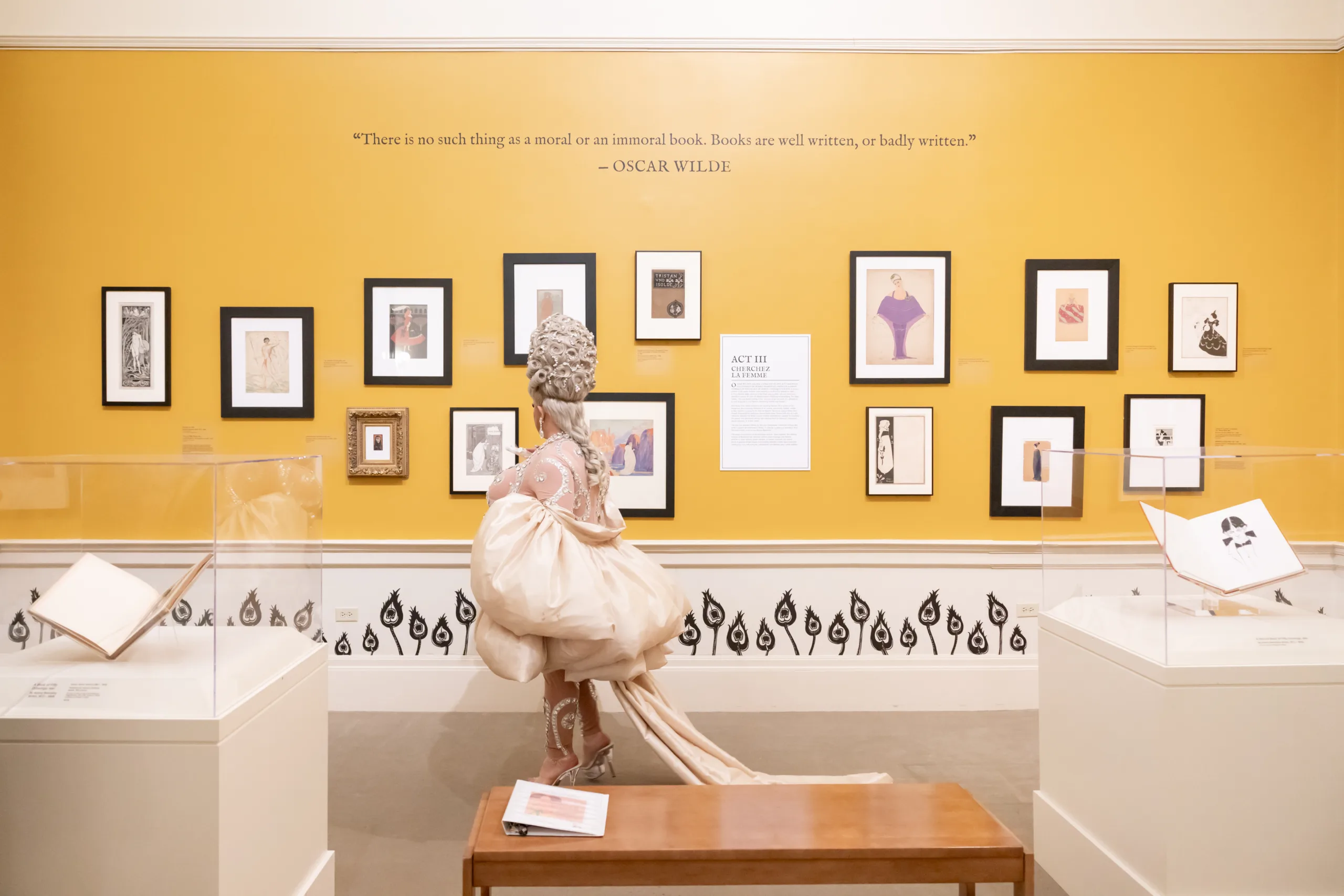 An art gallery with a yellow wall hung with small framed works. A patron dressed in a sumptuous 18th-century inspired outfit views the works.