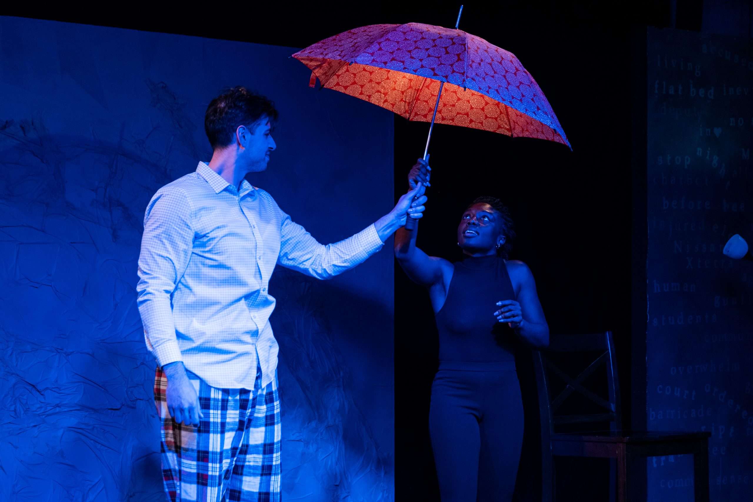 Two performers are on a stage lit with blue light. One holds an umbrella over the other.