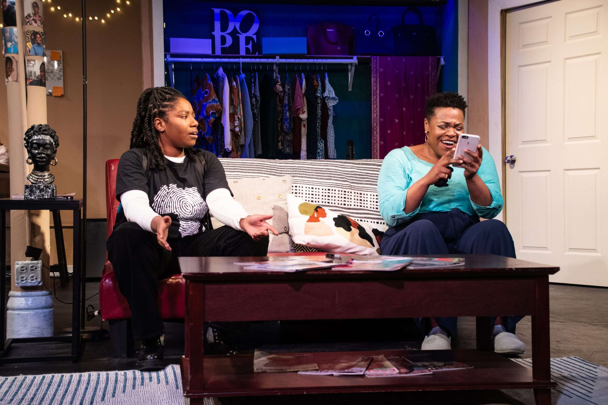 Two female actors seated on a couch in front of a coffee table. The actor on the right is holding a phone and the actor on the left is speaking.
