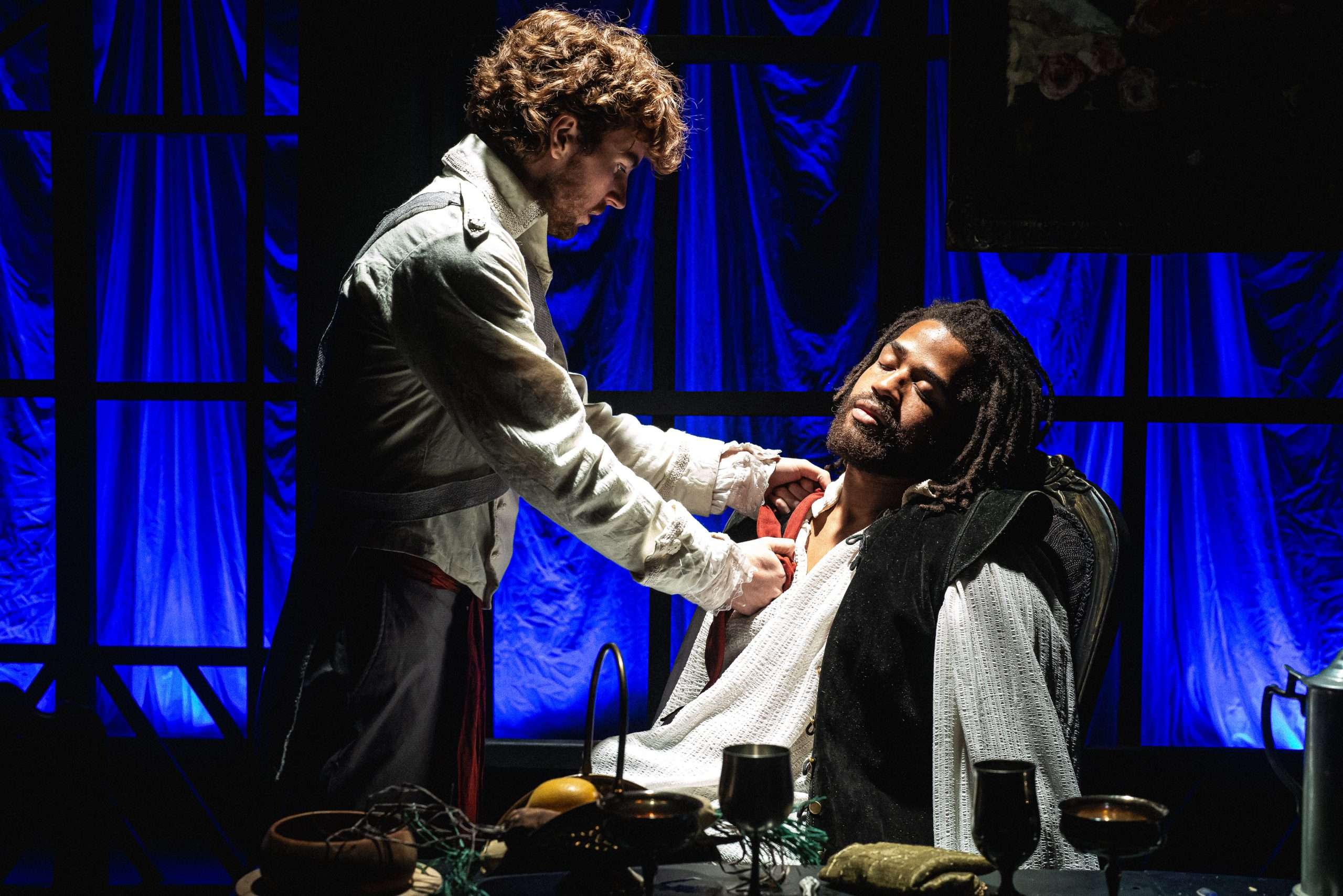 Two male actors are on stage with a blue curtain backdrop. One is seated with his eyes closed and the other is tending to him.