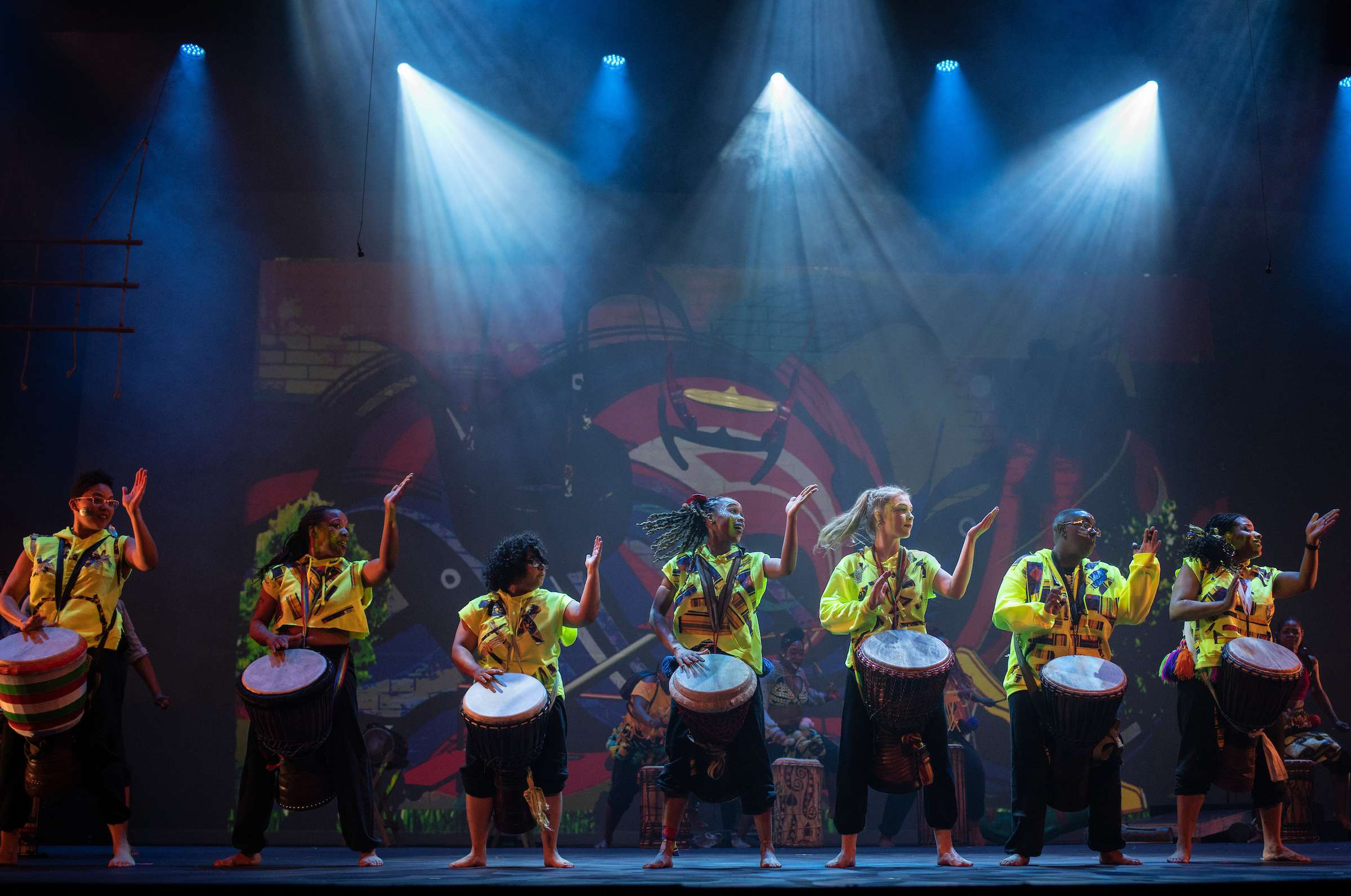 Seven women in a line dancing on stage with drums