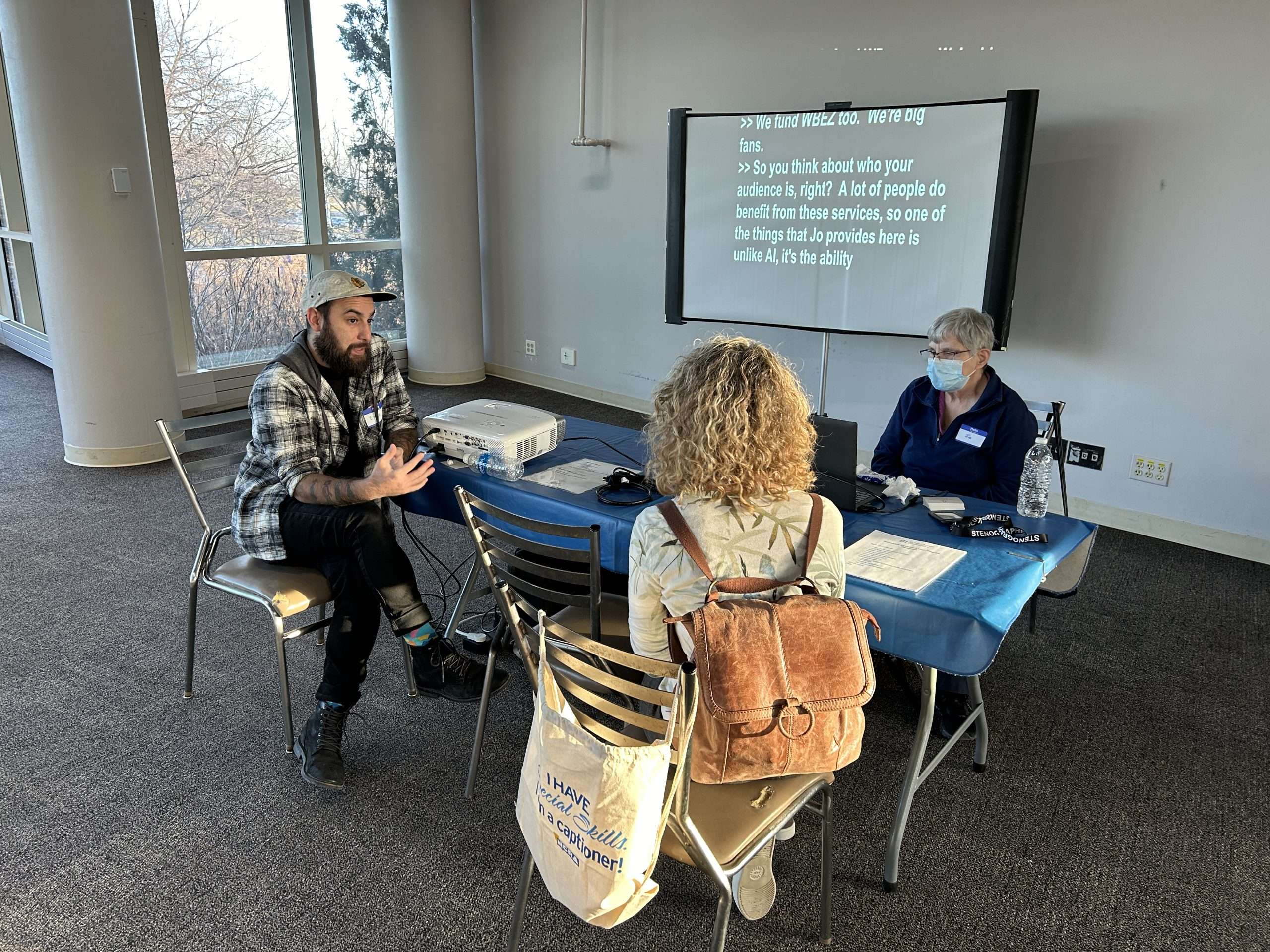 A man and two women are sitting at a table in front of a television with an audio transcript on it. One woman is wearing a mask and the other woman has a brown backpack on.