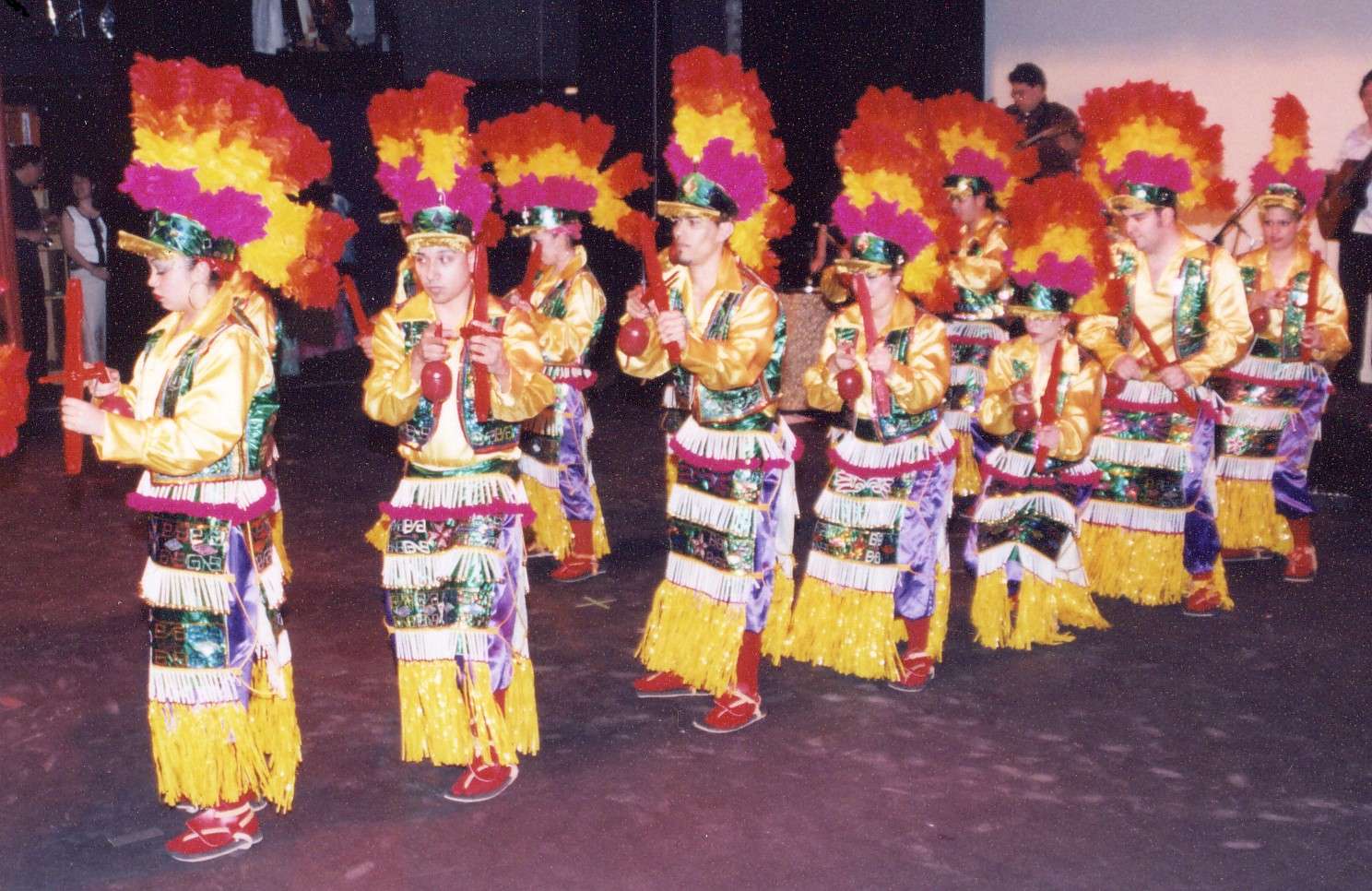 Nine dancers in red, pink, and yellow costumes are lined up holding instruments.