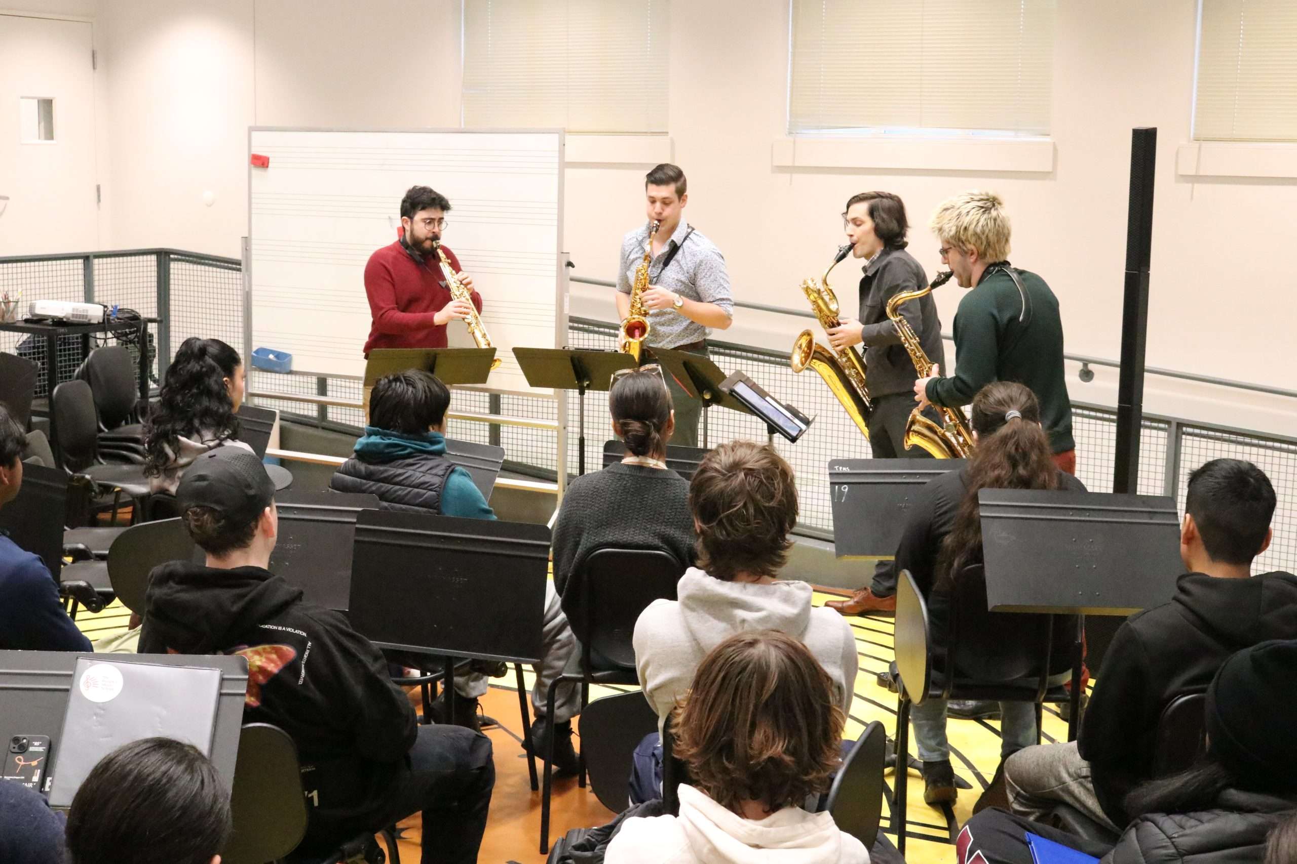 Four musicians playing saxophones in front of other musicians sitting down