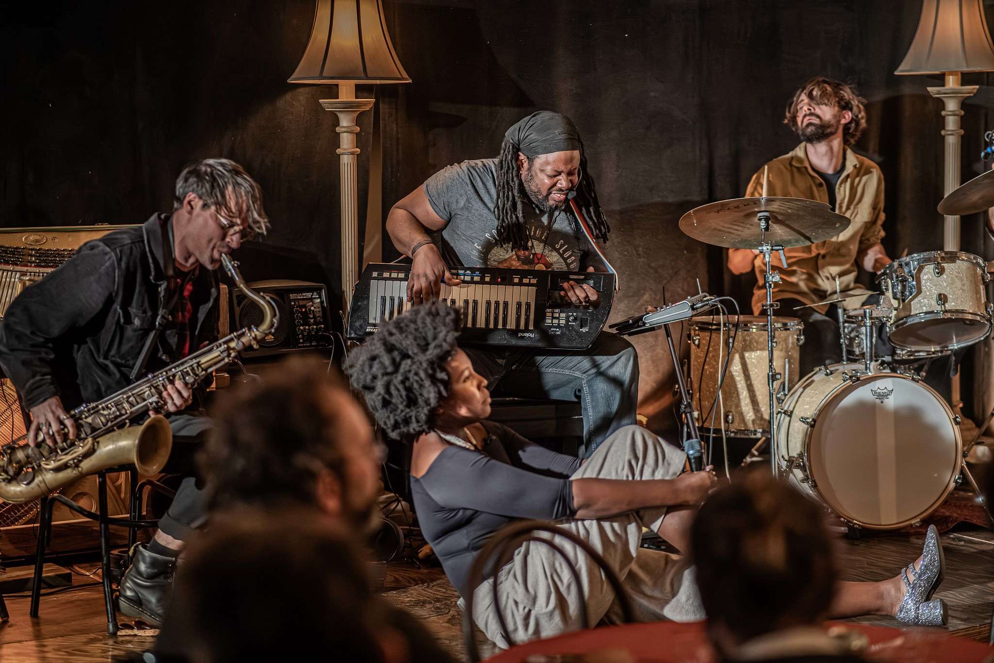 Three musicians are sitting down on stage playing instruments. One singer sits in front of them, holding a microphone. The audience is seated at tables, watching.