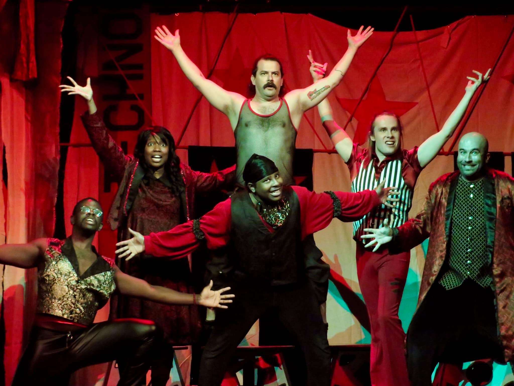 Six actors posing on stage wearing red and black costumes with a red backdrop